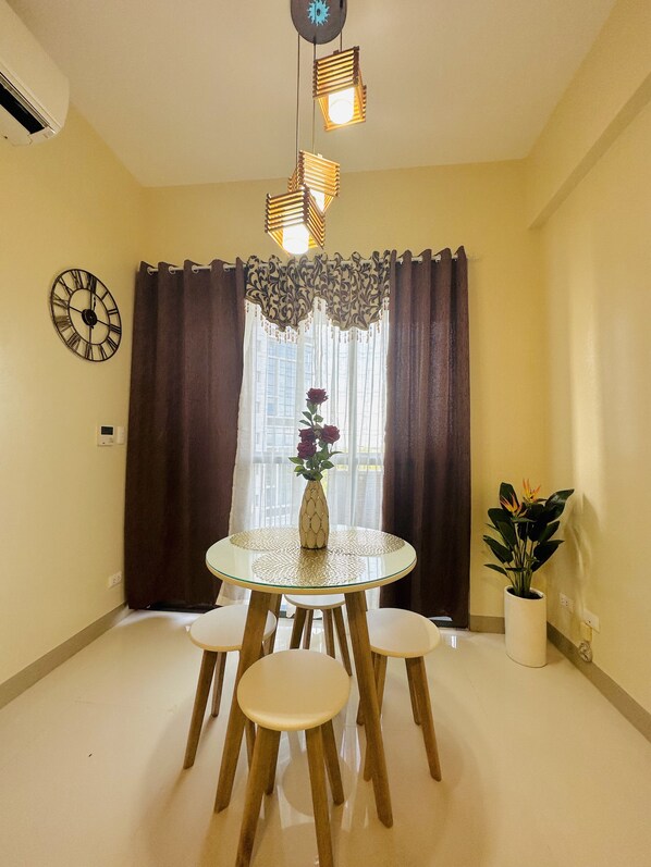 A dining area with a round table, four chairs, a vase with flowers, a wall clock, and a window with curtains.