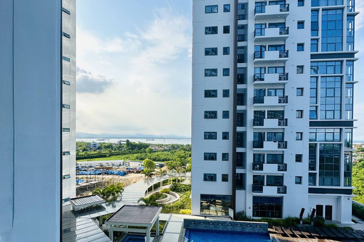 Un edificio de gran altura con piscina en la azotea y vistas a una zona ajardinada con árboles y un paisaje distante.