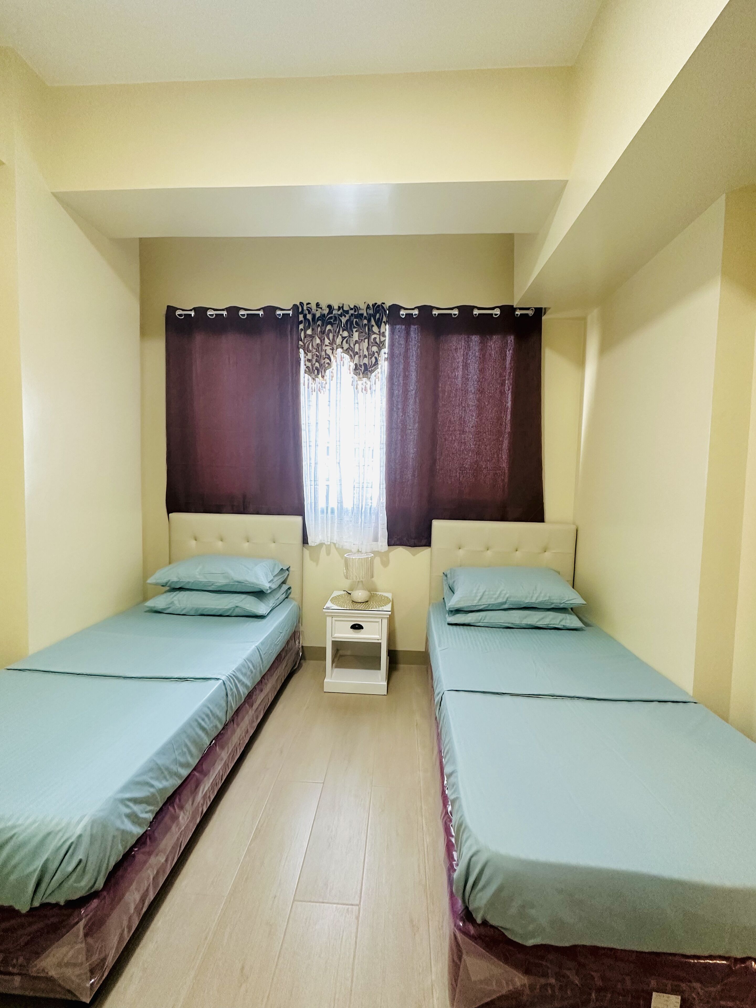 Une petite chambre propre, avec deux lits simples, une table de chevet, une lampe et une fenêtre avec des rideaux.
