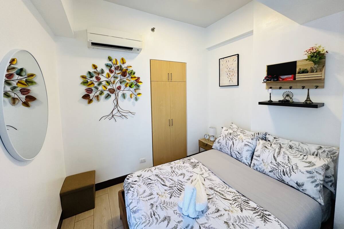 Un dormitorio con cama, armario, aire acondicionado de pared, una planta decorativa, un cuadro enmarcado y una estantería con objetos.