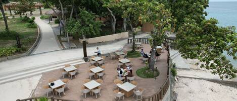 An outdoor dining area with tables and chairs, surrounded by trees and a view of the beach.