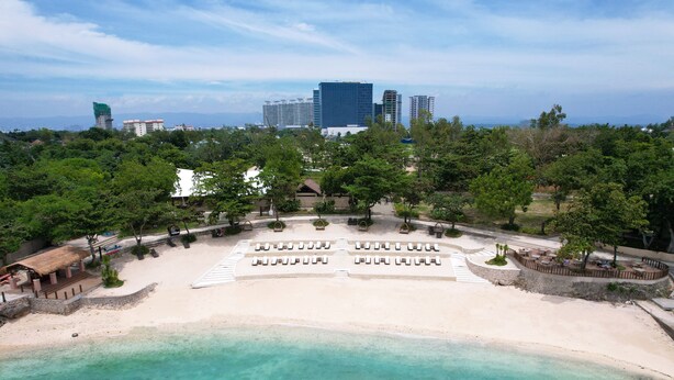 Un resort frente a la playa con arena, sillas de descanso y una vista del horizonte de la ciudad al fondo.