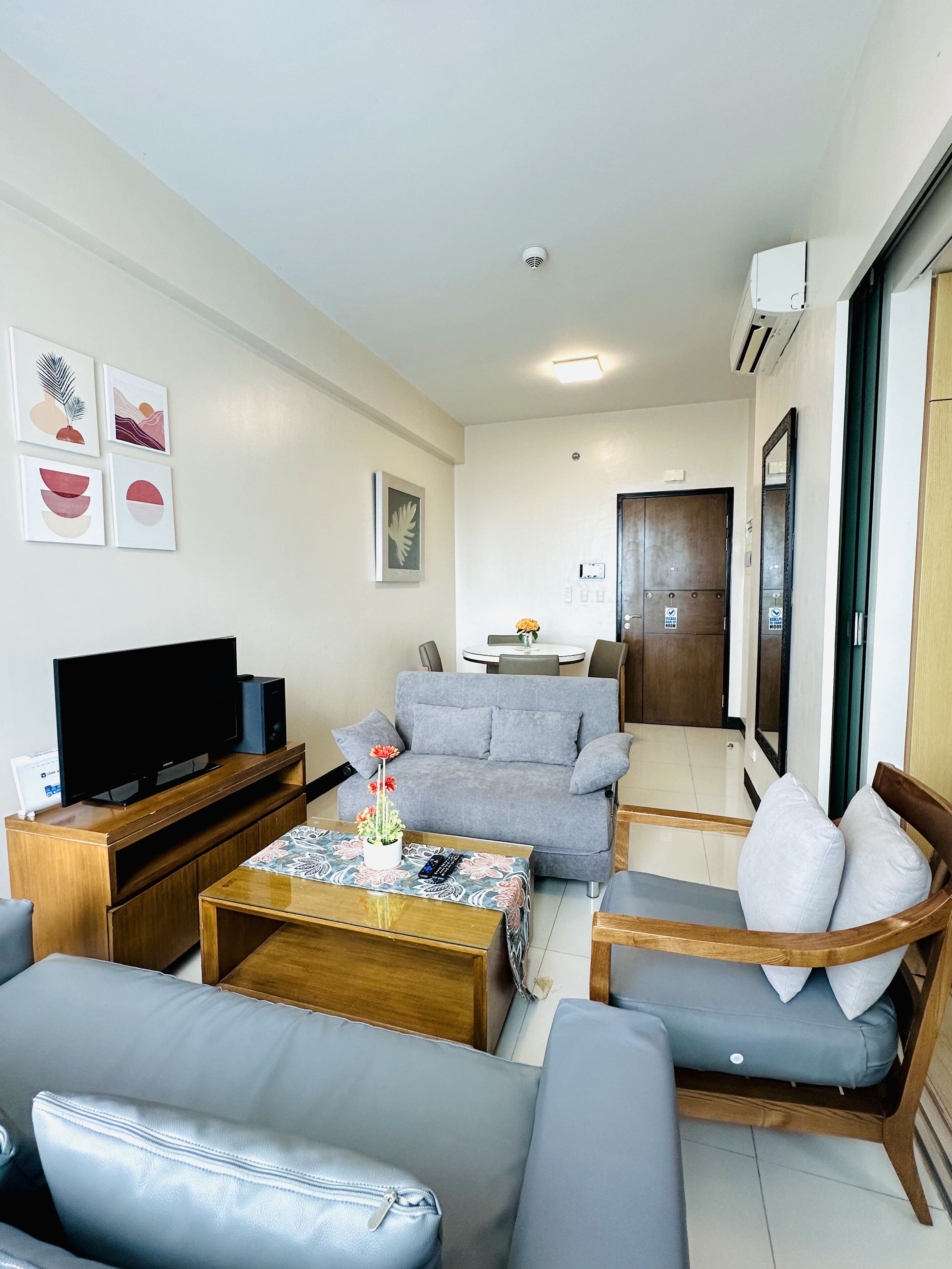 Una sala de estar moderna con un televisor de pantalla plana, sofás grises, una mesa de centro con un centro de mesa floral y un comedor al fondo.