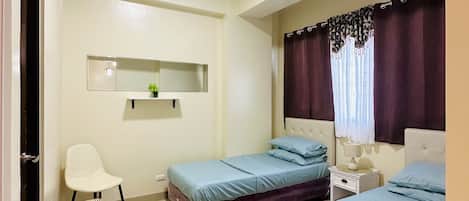 Un dormitorio con dos camas, una silla pequeña, una mesita de noche, un estante en la pared y una lámpara en el techo.