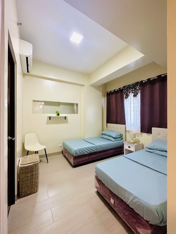 A bedroom with two beds, a small chair, a bedside table, a wall-mounted shelf, and a ceiling light.