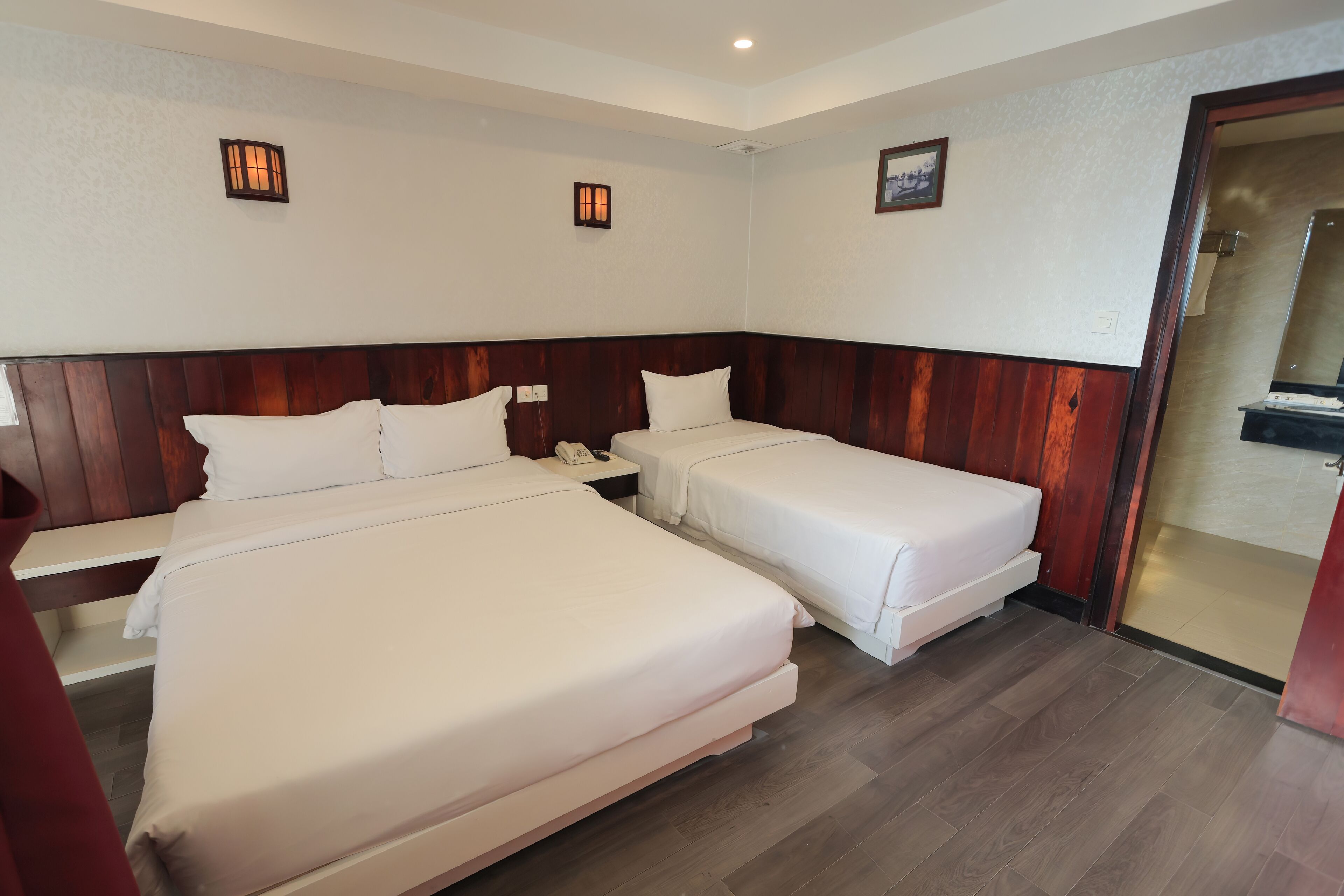 A hotel room with two beds, a wooden headboard, a bedside table with a phone, and a small wall-mounted light fixture.