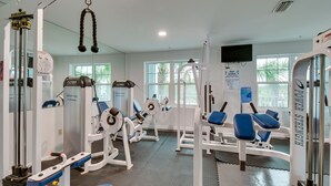 Ein Fitnessstudio mit verschiedenen Trainingsgeräten, einem an der Wand befestigten Fernseher und großen Fenstern, die viel Tageslicht hereinlassen.