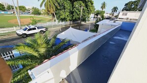 Vista de una calle con un coche blanco estacionado al lado, una carpa blanca y un área de piscina azul con palmeras.