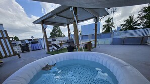 Una piscina inflable con un adorno de tiburón, una zona de estar cubierta y un patio vallado con sillas de descanso y una parrilla.