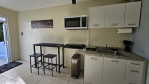 Cocina compacta con microondas, lavamanos y alacenas. Hay una pequeña mesa con dos taburetes y un ventilador de techo.