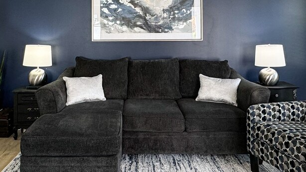 A dark blue room with a grey sofa, two matching side tables, and a framed abstract painting on the wall.