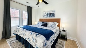 Un dormitorio con cama, ventilador de techo, mesita de noche, lámpara y ventana con cortinas.
