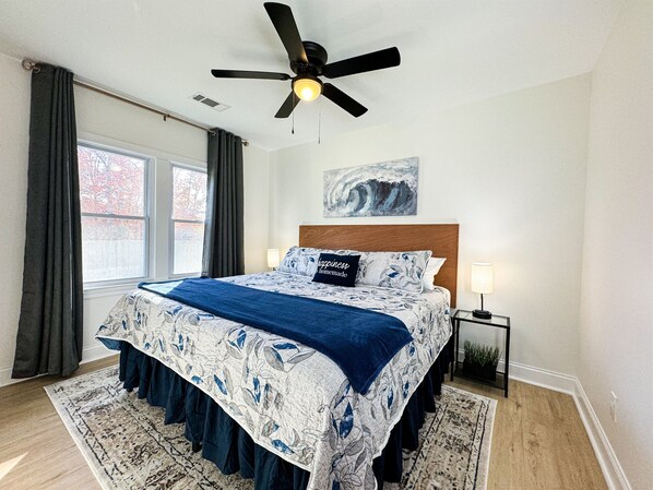 Un dormitorio con cama, ventilador de techo, mesita de noche, lámpara y ventana con cortinas.