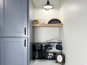 Een keukenhoek met een koffiezetapparaat, een rek voor koffiemokken en een bordje met de tekst 'Our Little Coffee Bar'.
