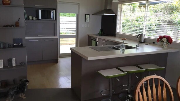 A modern kitchen with a central island, bar stools, and a view of greenery outside. - Pacific Vistas (Athenree)