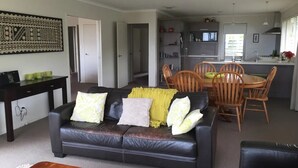 A modern living room with a black leather sofa, wooden dining table, and chairs. There is a kitchen area with a sink and cabinets in the background. - Pacific Vistas (Athenree)