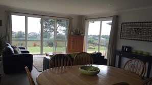 A dining area with a round wooden table, four chairs, and a green bowl. There is a wooden cabinet and a sofa with cushions. Large windows provide a view of the outdoors. - Pacific Vistas (Athenree)