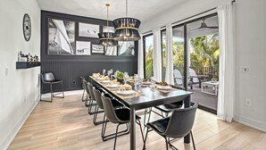 Een eetruimte met een lange tafel gedekt voor een maaltijd, omringd door stoelen, met uitzicht op een balkon en palmbomen.