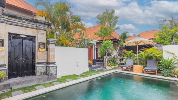 Una piscina con sillas de descanso, un edificio de piedra con una puerta negra y plantas tropicales.