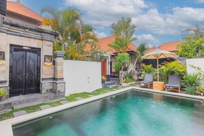 Una piscina con sillas de descanso, un edificio de piedra con una puerta negra y plantas tropicales.