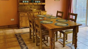 A dining area with a wooden table set for four, surrounded by chairs. A wooden cabinet with shelves is against the wall, and a window with curtains is visible. - Sunshine Heights by Interhome (Kenmare)