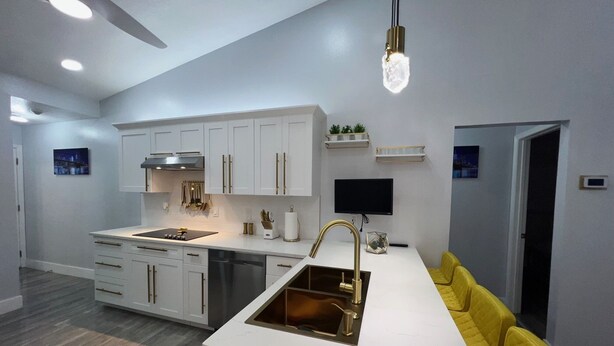 Una cocina moderna con armarios blancos, horno empotrado y una isla grande con pileta. Hay un sofá amarillo y un televisor de pantalla plana montado en la pared.