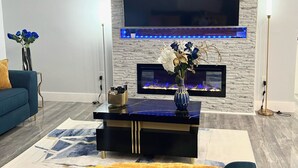 Una sala de estar moderna con una televisión de pantalla plana montada en una pared con textura, una chimenea con luz LED azul, un sofá azul y un jarrón decorativo sobre una mesa de centro negra.