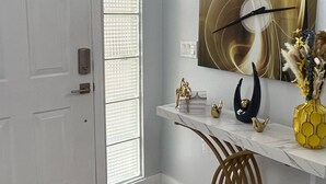 Un pasillo con una puerta blanca, un estante de mármol decorado con objetos y un reloj de pared, además de un ventanal con persianas.