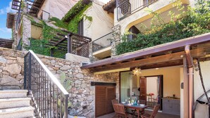 Un escalier en pierre mène à une terrasse en bois aménagée avec du mobilier d’extérieur, un mur en pierre et un bâtiment doté de balcons et de verdure.
