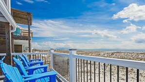 Eine Holzterrasse mit blauen Stühlen und einem weißen Geländer mit Blick auf einen Strand, Dünen und das Meer.