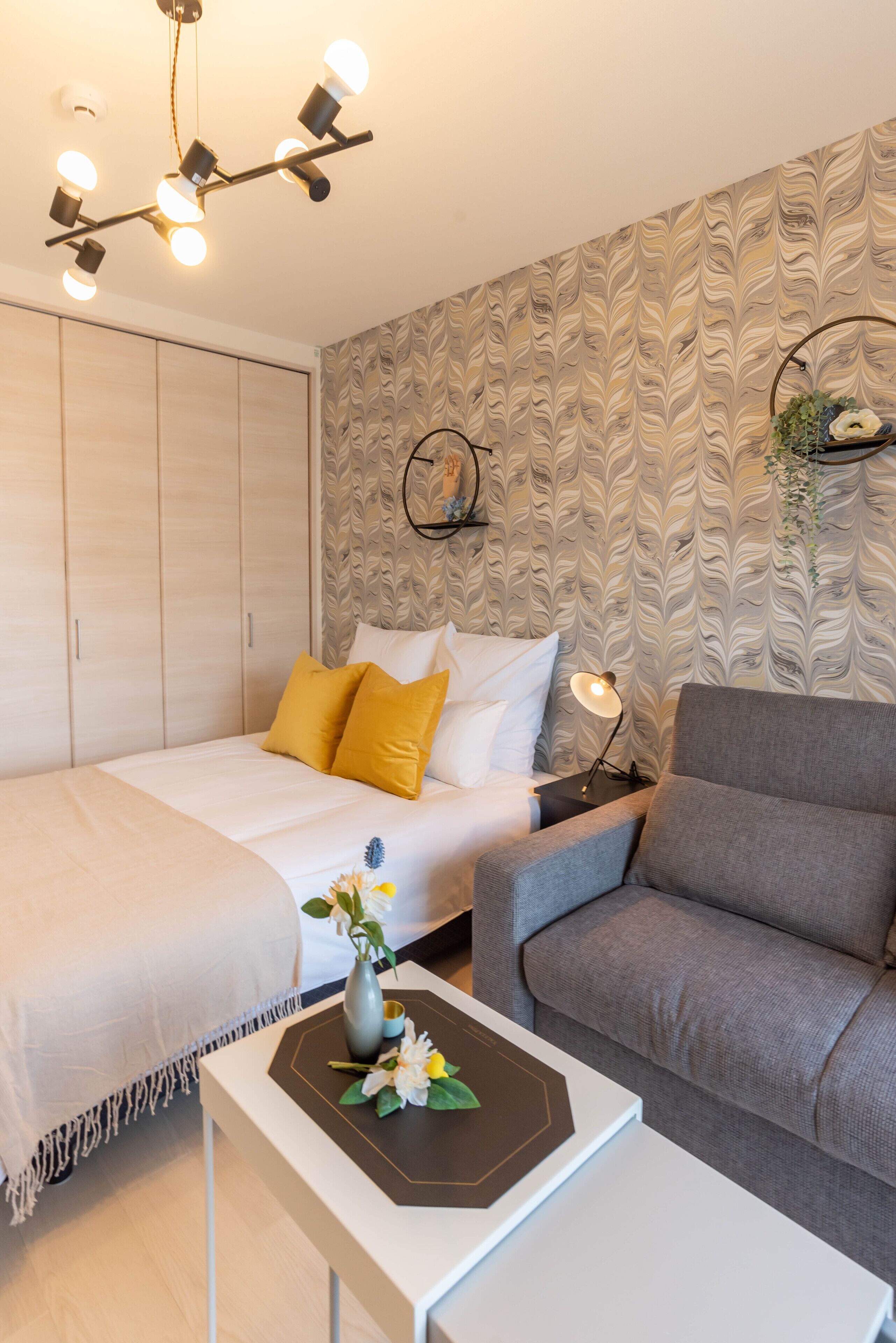 Un dormitorio moderno con cama, un sofá gris, una mesita de noche con un florero y un papel tapiz estampado.