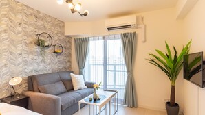 Ein modernes Hotelzimmer mit grauem Sofa, einem kleinen weißen Tisch, einer Topfpflanze und einem großen Fenster mit Vorhängen.