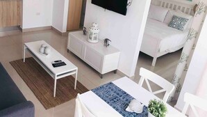 A modern, well-lit room with a white sofa, a dining table with chairs, a white cabinet with decorative items, a flat-screen TV mounted on the wall, and a bed with a white headboard.