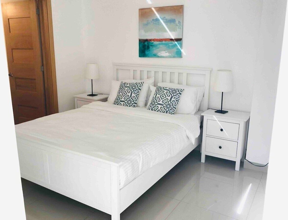 A white bed with patterned pillows, a nightstand with a lamp, and a painting on the wall.