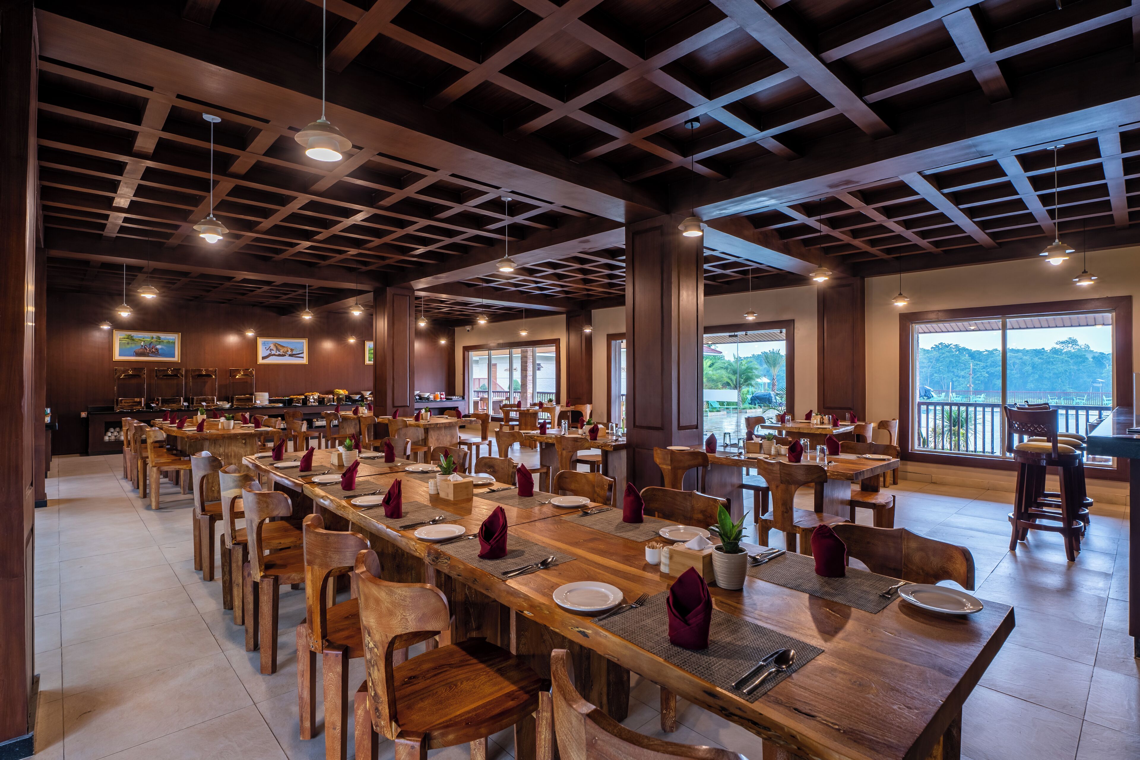 A spacious dining area with long wooden tables and chairs, large windows offering views of the outdoors, and a high ceiling with exposed beams.