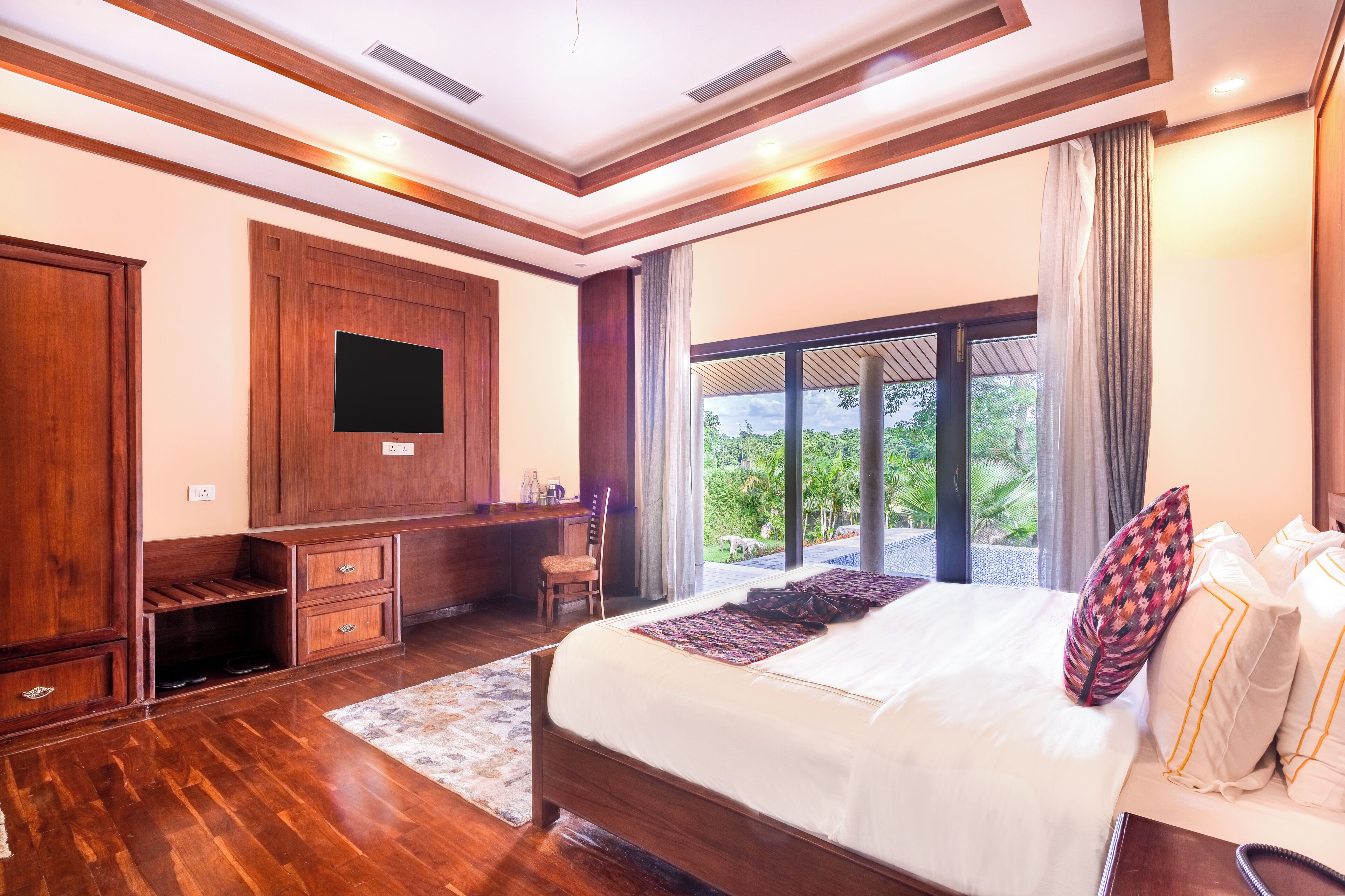 Un dormitorio amplio con una cama grande, muebles de madera, un televisor de pantalla plana y un balcón con vistas a zonas verdes.