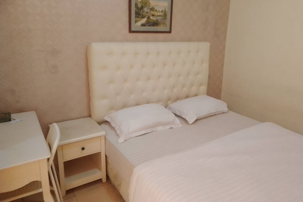 A neatly arranged bedroom with a bed, two pillows, a bedside table, and a small drawer. A framed picture hangs on the wall above the bed.