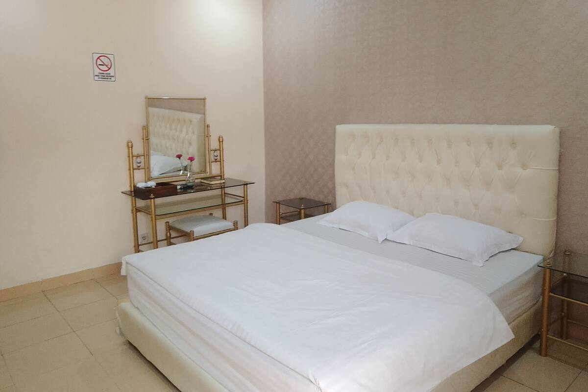 A hotel room with a large bed, a bedside table, a mirror, and a small desk with a chair.