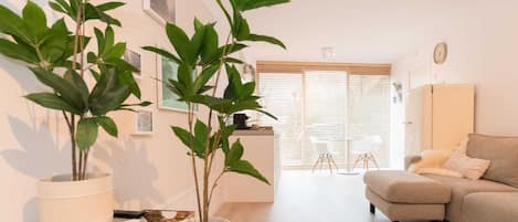 A modern living room with a sofa, a coffee table, and two potted plants. There is a dining area with a chair and a table in the background.
