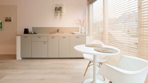 A modern kitchen with a white countertop, a sink, and a window with blinds. There is a dining area with a white table and chairs. - Studio Loet Schagen (Schagen)