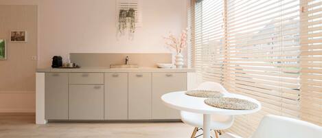 Una cocina moderna con encimera blanca, un lavamanos y un ventanal con persianas. Hay una zona de comedor con una mesa y sillas blancas.