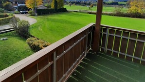 Un balcon donnant sur un champ verdoyant, un bâtiment et des montagnes au loin.