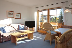 A living room with a sofa, a wooden coffee table, wicker chairs, a television, and a large window with a view of trees. - Apartment 'Ferienwohnung Hörnerblick 32' with Mountain View, Balcony and Wi-Fi (Sonthofen-Altstädten)