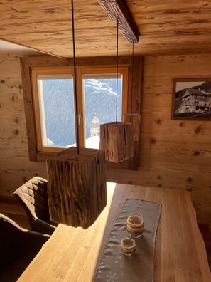 A wooden cabin interior with a dining table, chairs, and a view of snow-capped mountains through a window.