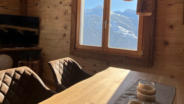 Un intérieur en bois comprenant une table à manger, des chaises, un téléviseur et une fenêtre donnant sur la montagne.