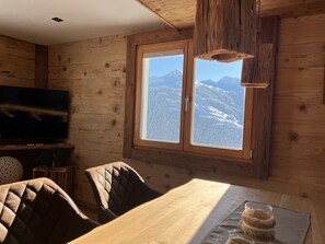 Ein Holzinterieur mit Esstisch, Stühlen, einem Fernseher und einem Fenster mit Blick auf die Berge.