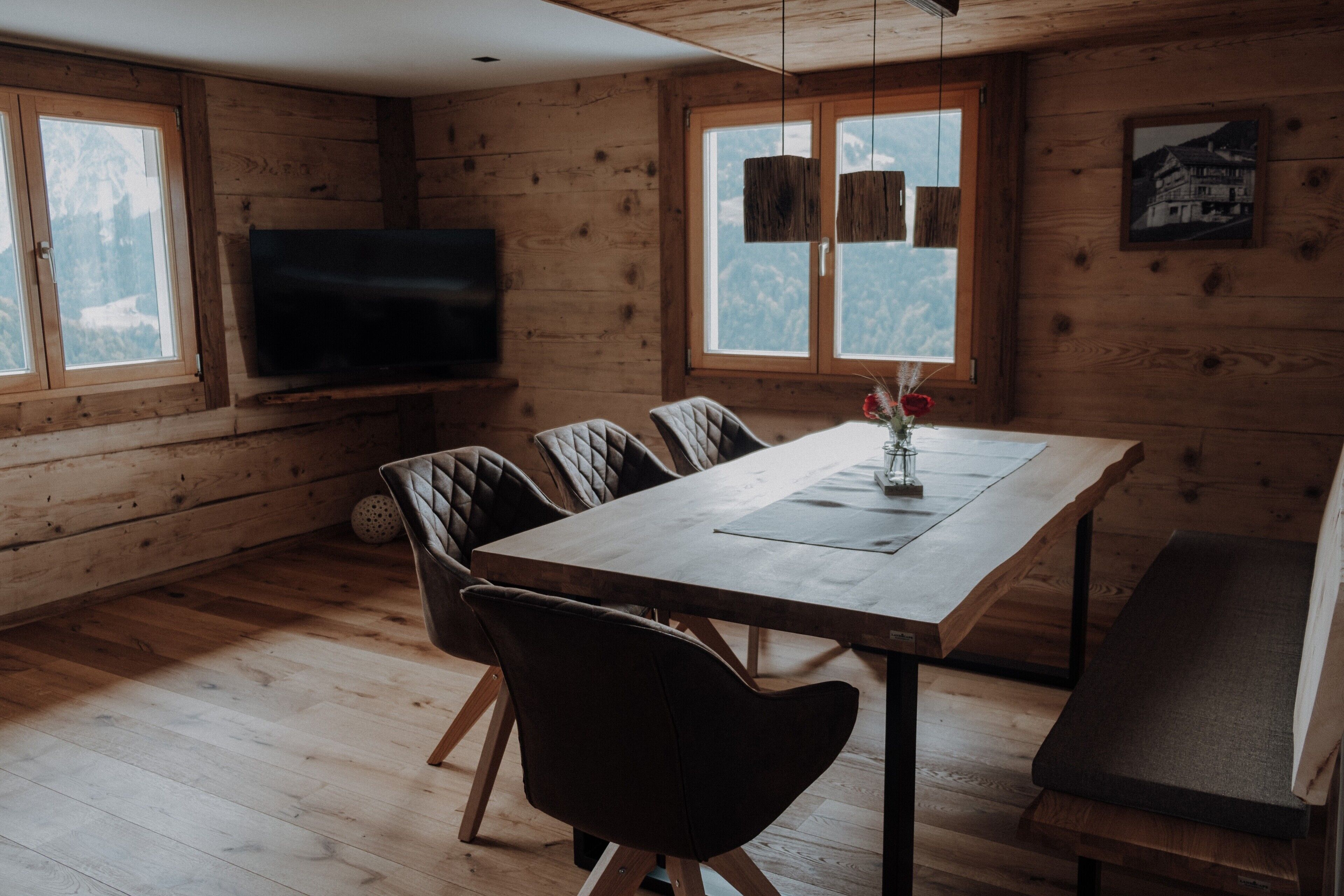 A wooden interior with a dining table, chairs, a bench, a TV, and windows with a mountain view.