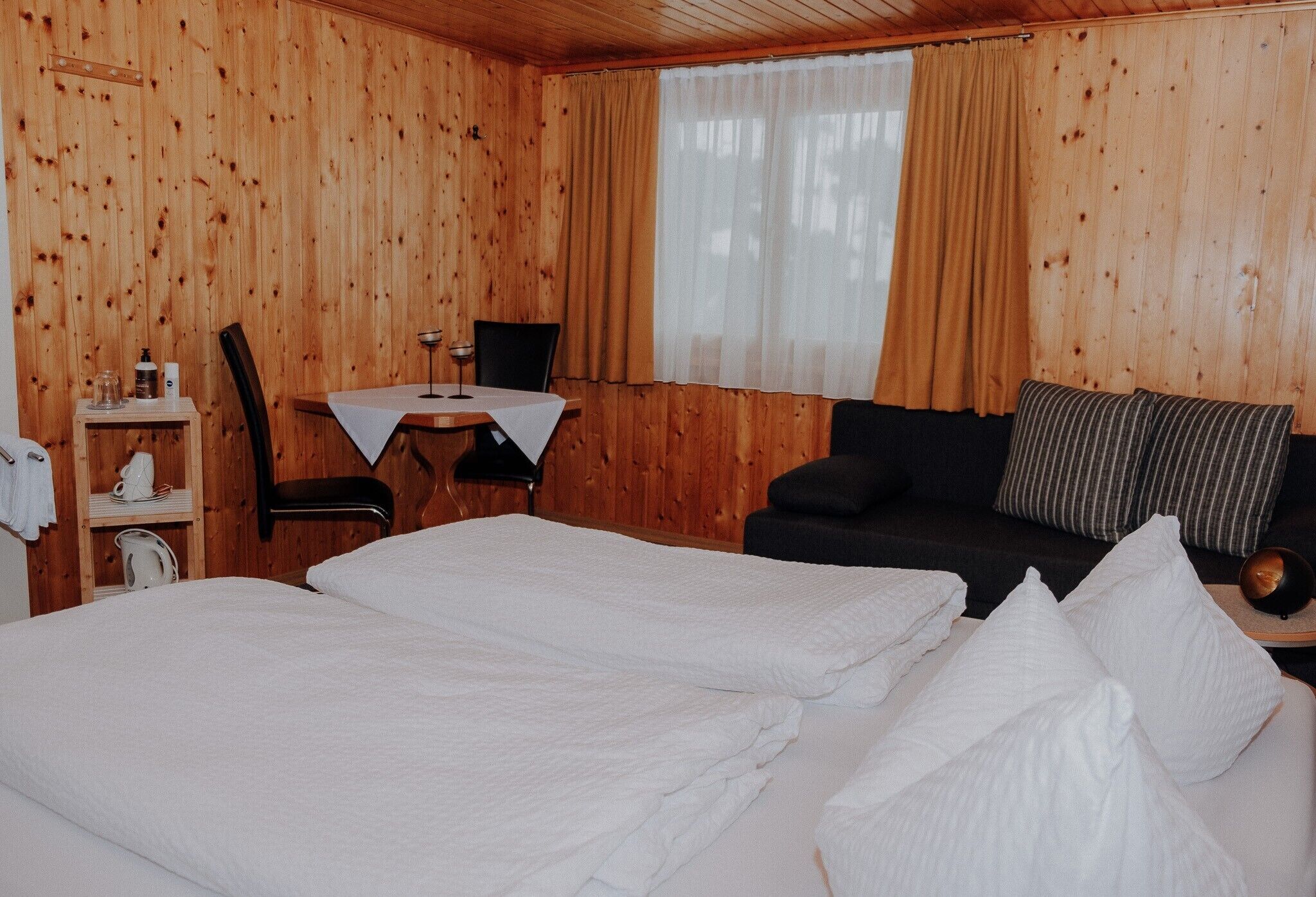 A wooden-paneled room with a bed, a small table with a chair, and a sofa with pillows.