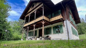 Une maison traditionnelle en bois, dotée d’un balcon et entourée de verdure.