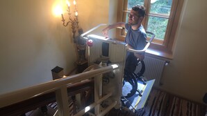 Une personne utilise un monte-escalier dans une pièce où se trouvent un radiateur, une fenêtre donnant sur un espace verdoyant et un chandelier avec des bougies allumées.